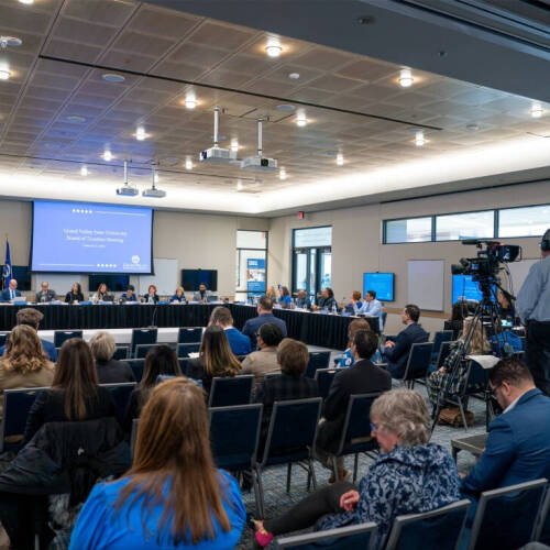 A wide-angle view of a group of people in a meeting held in a large, modern conference room. Some are seated along U-shaped table at the front, facing the rest. A large projection screen displays in the background.